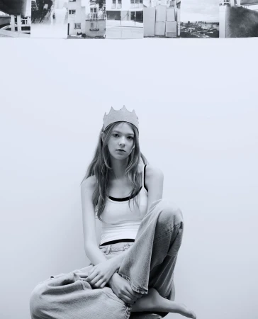 Black and white portrait of a young woman with crown. She sits on stool in front of a white wall, with architectural collage above.
