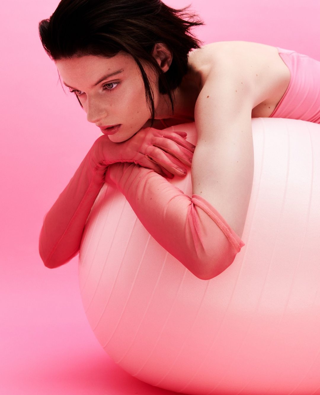 An adult female poses in a photo studio, lying on a pink exercise ball against a pink seamless backdrop, wearing a pink top and gloves.