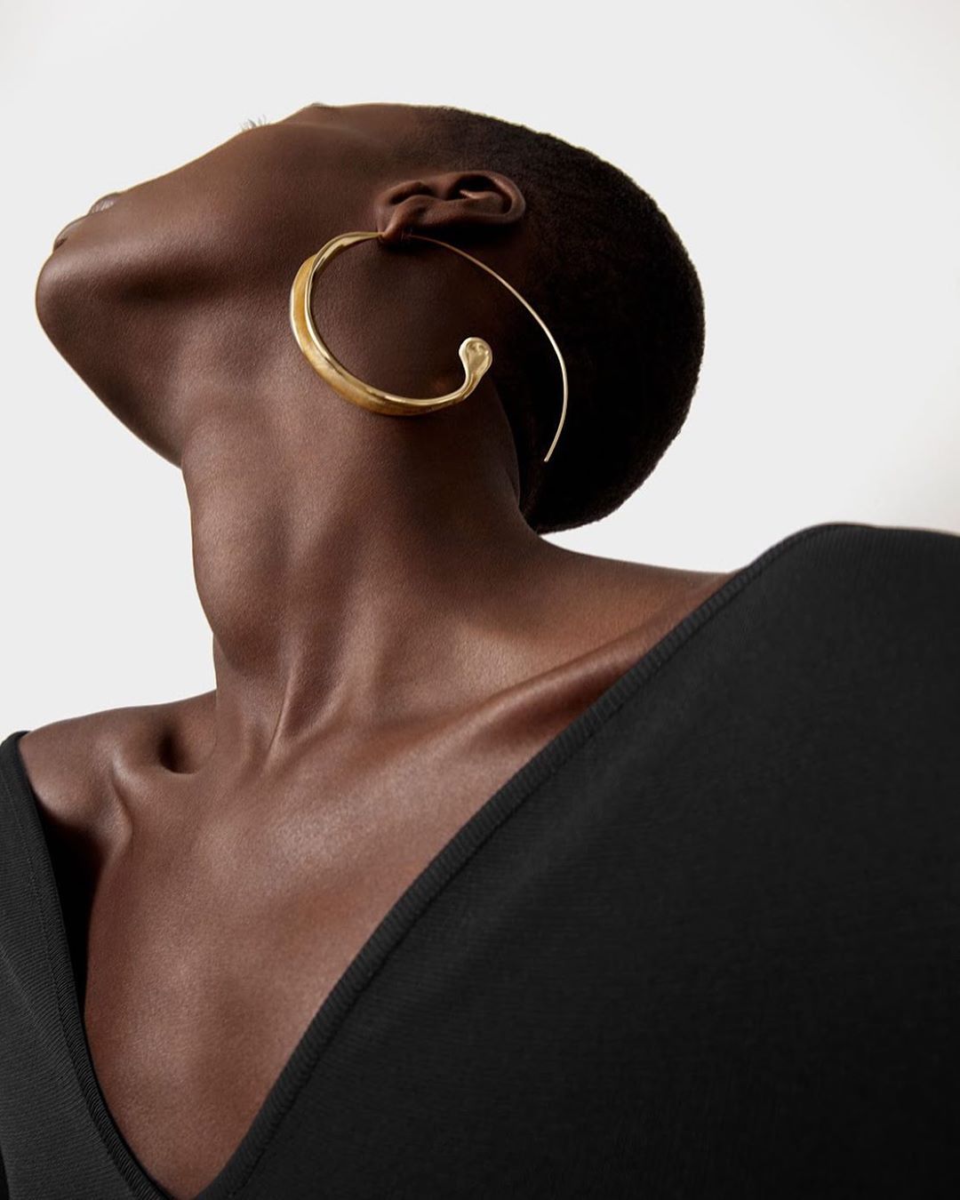 Portrait of dark-skinned adult woman looking up, wearing a black top and gold hoop earrings, against a neutral-toned background.