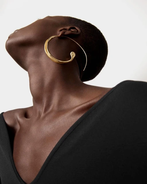 Portrait of dark-skinned adult woman looking up, wearing a black top and gold hoop earrings, against a neutral-toned background.