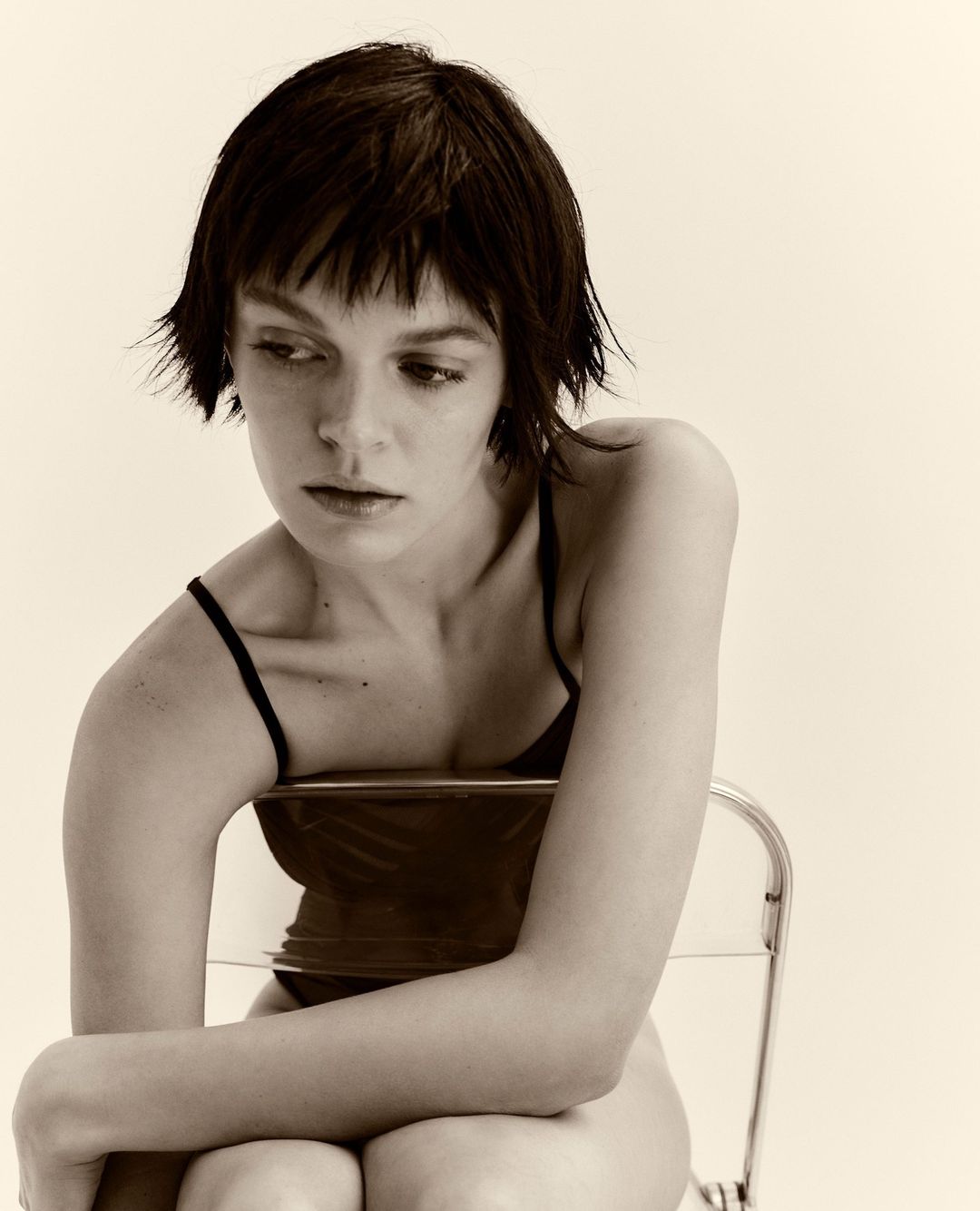 Portrait of a woman with dark short hair seated on a transparent chair against a pale background by Bibi Cornejo Borthwick