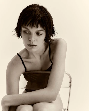 Portrait of a woman with dark short hair seated on a transparent chair against a pale background by Bibi Cornejo Borthwick