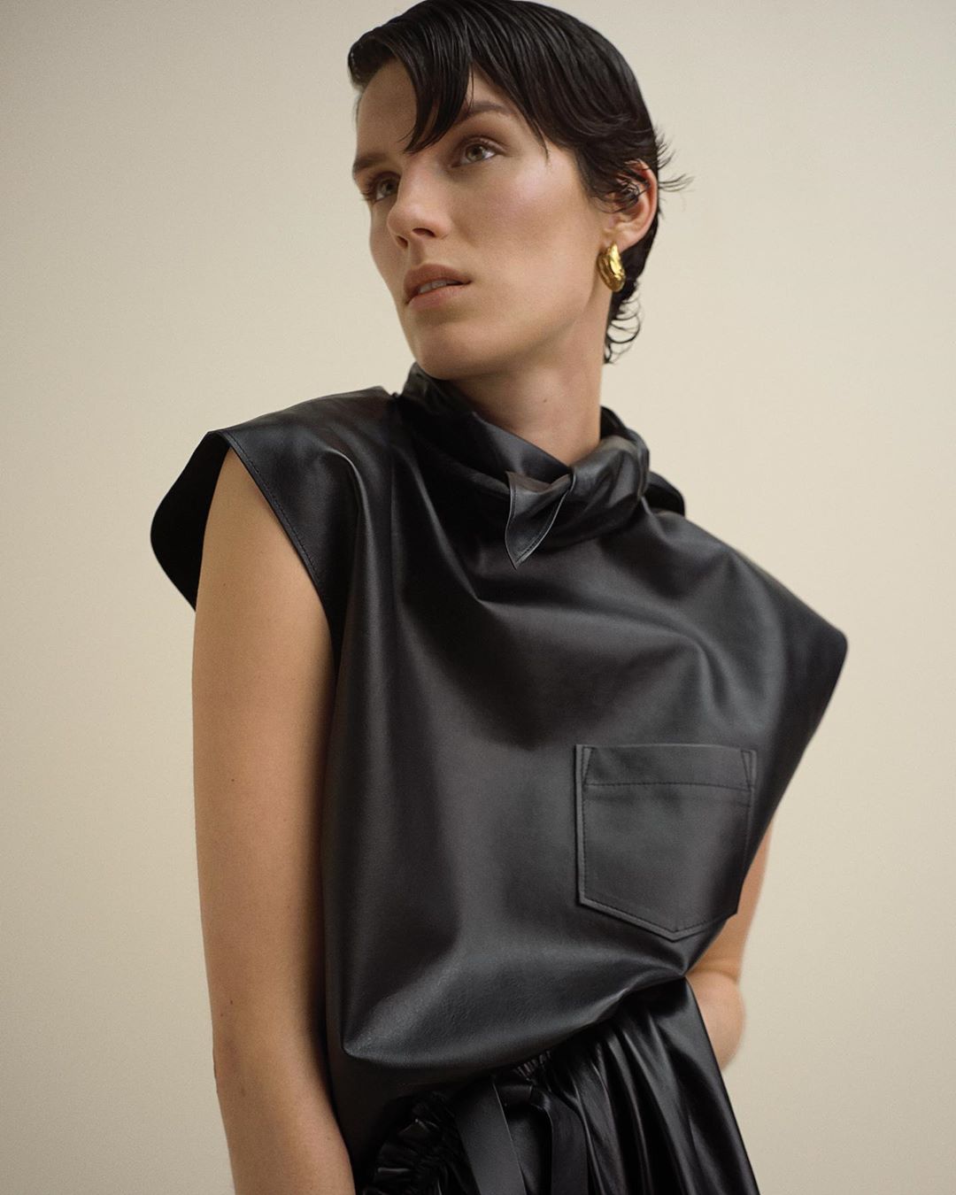 Portrait of a woman wearing a black sleeveless leather top and gold earrings against a plain light wall.