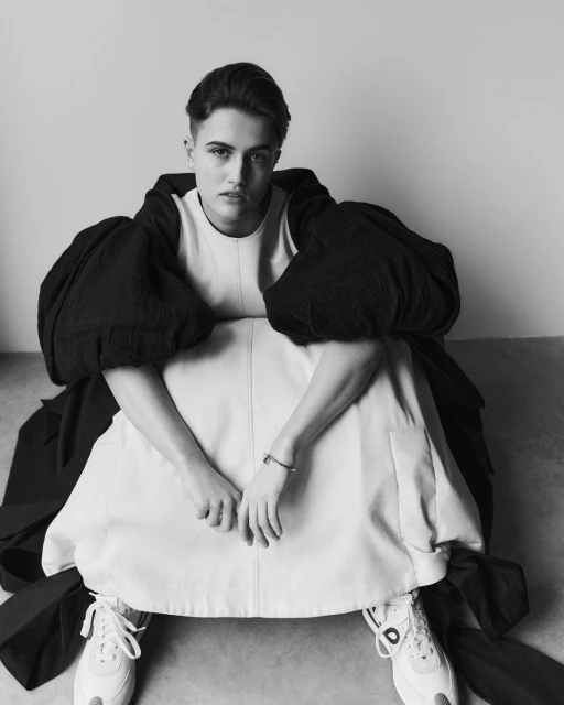 Black and white portrait of a model in a runway look with a large, black puffed sleeves and white dress sitting in front of a wall.