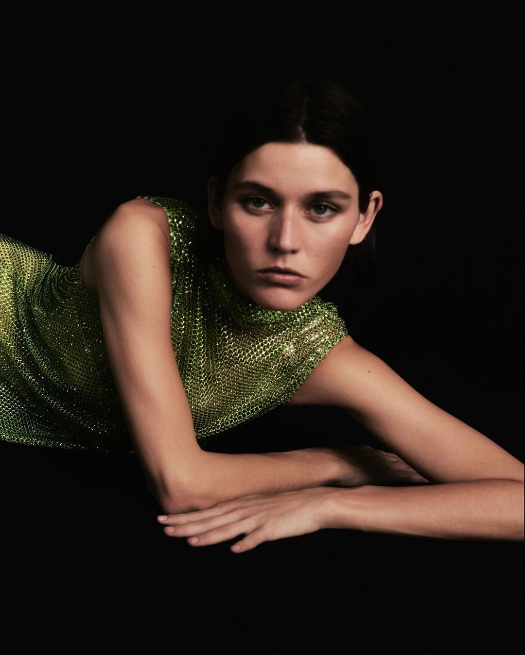 Woman in a green, chainmail dress lies on a black surface, posing for a fashion shot with a neutral facial expression in a studio.