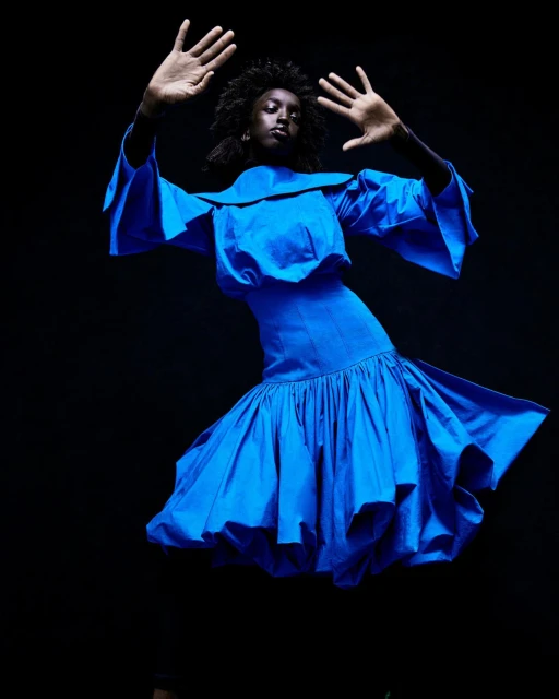 Fashion portrait of an African-American woman in a bright blue dress, posing with her hands up against a dark background in a photo studio.