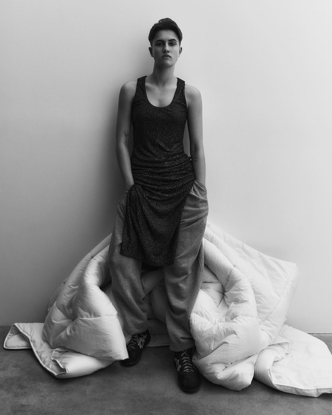 Monochrome portrait of a young adult woman in a sleeveless dress, baggy sweatpants, and sneakers, seated on a white comforter indoors.