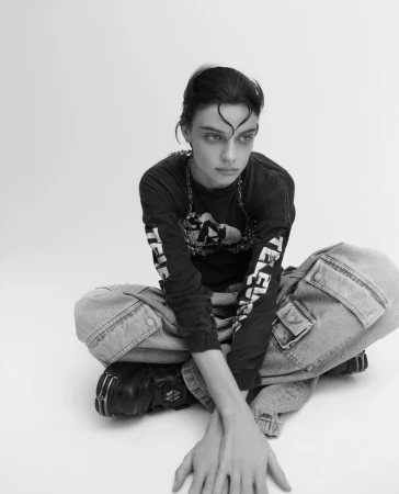 Black and white shot of a young adult woman with dark hair in a long-sleeve shirt and baggy jeans sitting cross-legged on a white surface.