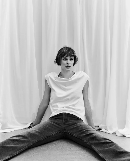 Studio shot of a woman in jeans and a white top sitting on the floor in front of a white backdrop in a relaxed pose.