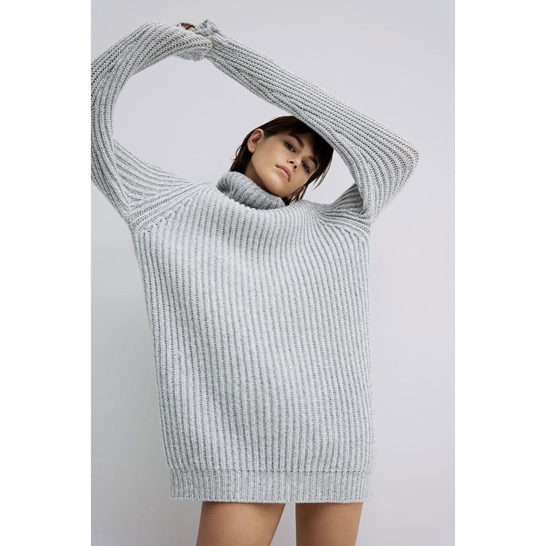 Woman wearing a grey ribbed-knit turtleneck sweater with arms raised, medium shot against a neutral grey studio background.