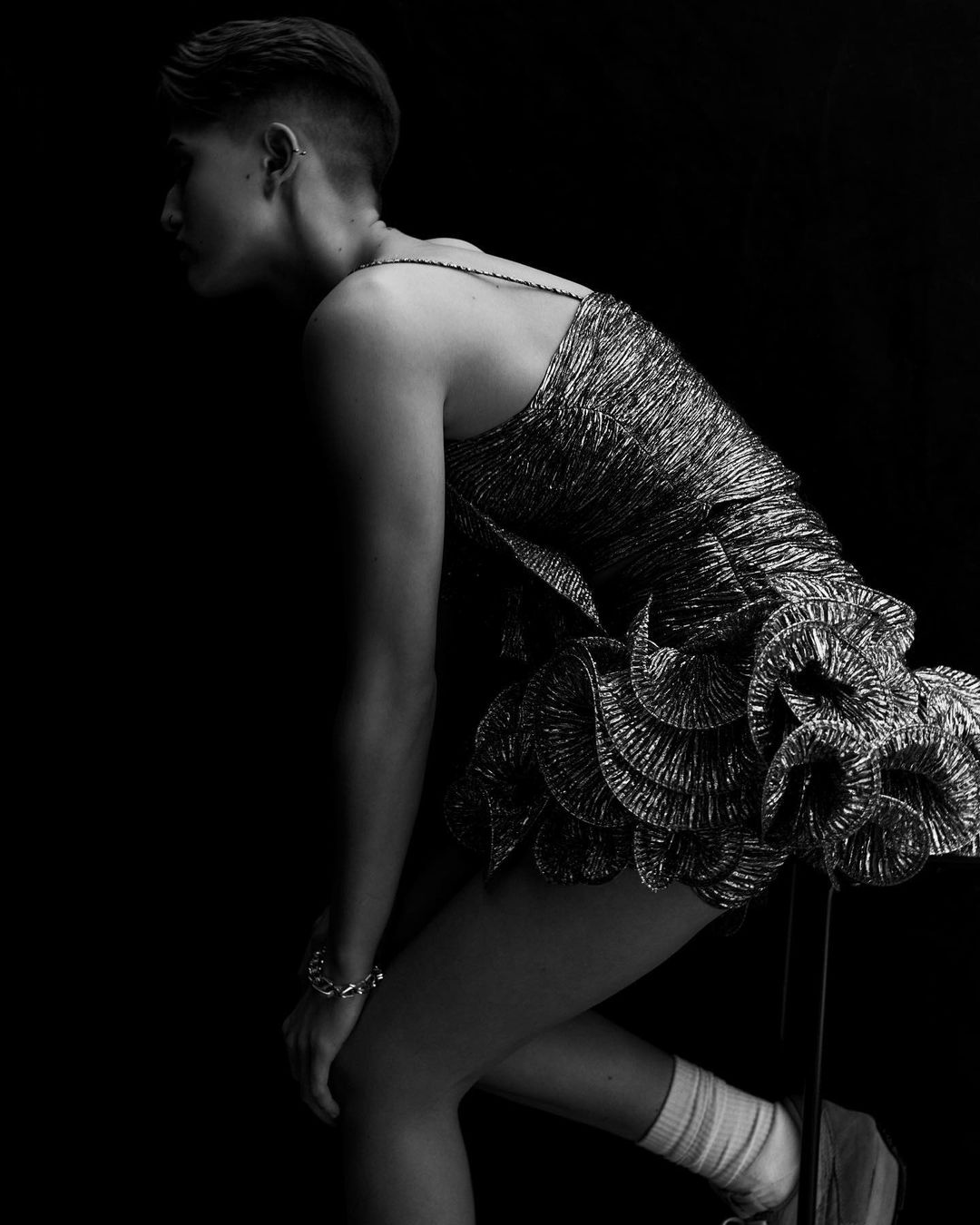 Black-and-white studio shot of a woman in a metallic dress, leaning forward with one knee bent, against a dark background.
