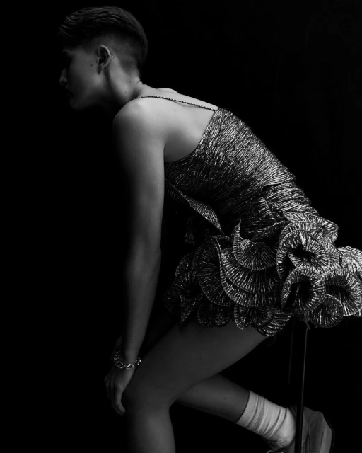 Black-and-white studio shot of a woman in a metallic dress, leaning forward with one knee bent, against a dark background.