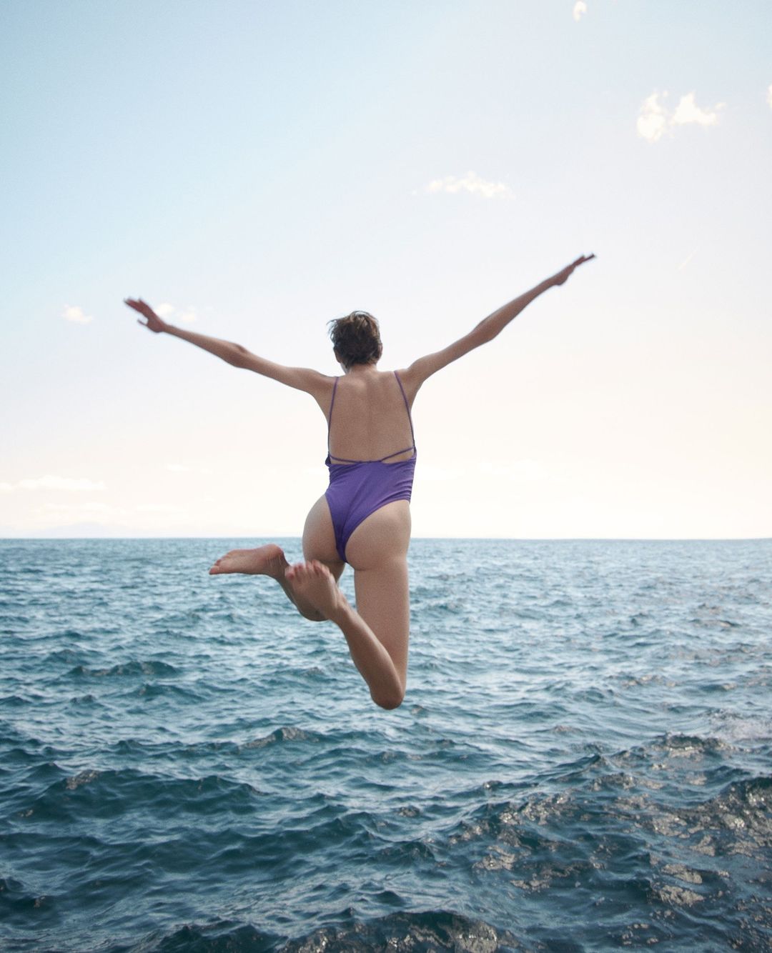 Woman in purple swimsuit leaps into blue ocean water against a sunny sky, arms outstretched. Summer, travel, and watersports.