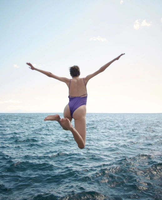 Woman in purple swimsuit leaps into blue ocean water against a sunny sky, arms outstretched. Summer, travel, and watersports.