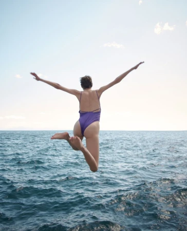 Woman in purple swimsuit leaps into blue ocean water against a sunny sky, arms outstretched. Summer, travel, and watersports.