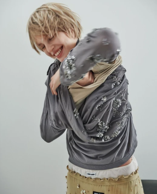 Smiling young adult woman adjusts a grey, embellished long-sleeve shirt over a tan top, complemented by a tan skirt with Miu Miu logo.