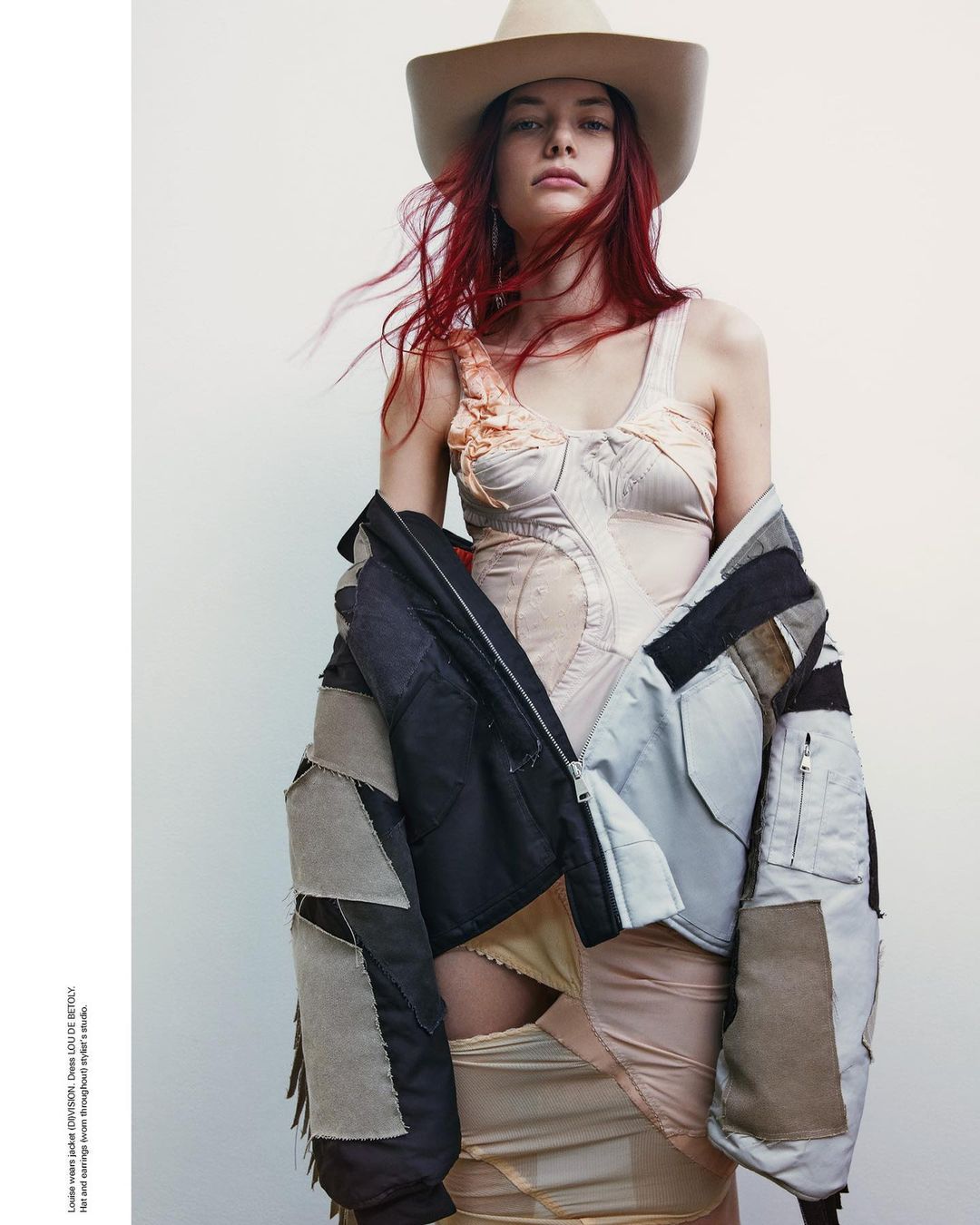 A woman with red hair and a cowboy hat poses in a light-colored corset top and a patchwork jacket against a light background.