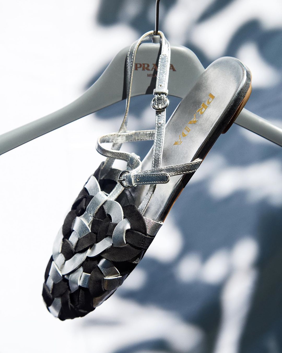 Prada sandal on a hanger, showcasing a black and silver woven design, displayed against a soft, blurred background with ambient lighting.