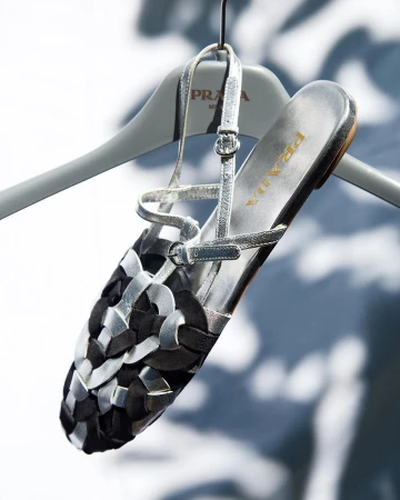 Prada sandal on a hanger, showcasing a black and silver woven design, displayed against a soft, blurred background with ambient lighting.