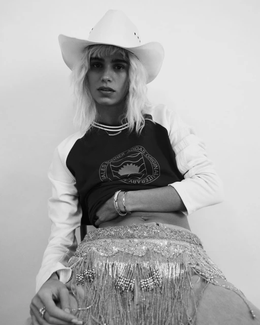 Monochrome portrait of a person wearing a cowboy hat, a t-shirt with long sleeves and a beaded belt with fringe against a plain background.