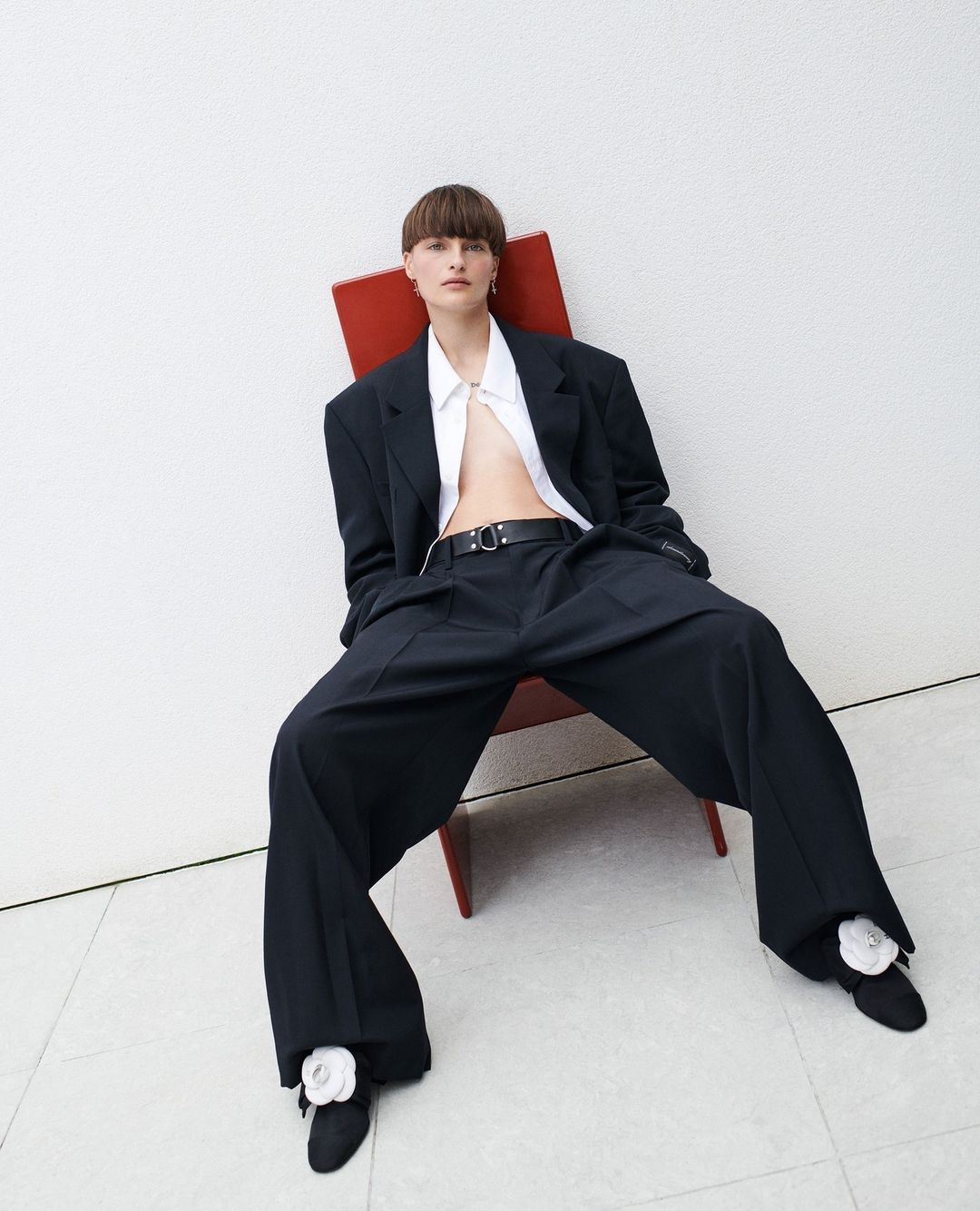 Elegant female fashion model sits in a red chair with oversized black suit and pants, white button shirt, short brown hair in studio.