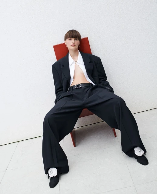 Elegant female fashion model sits in a red chair with oversized black suit and pants, white button shirt, short brown hair in studio.
