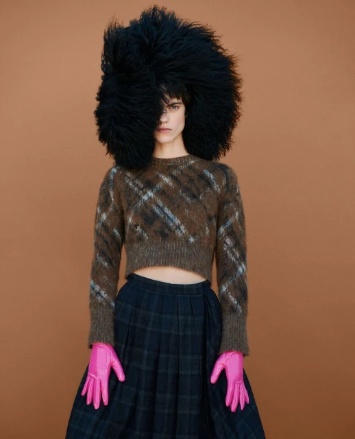 Female model wearing a brown plaid sweater with a black skirt and a large, dark, shaggy hat against a neutral brown background.