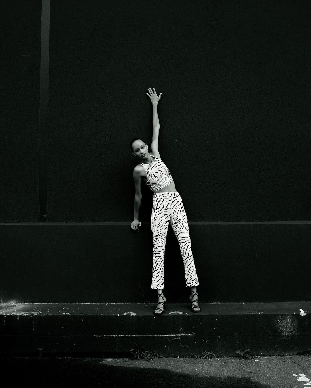 Monochrome shot of a solo adult female model in a zebra-print outfit leaning on a black wall with one arm raised above her head.
