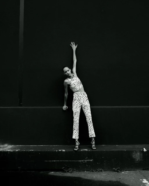 Monochrome shot of a solo adult female model in a zebra-print outfit leaning on a black wall with one arm raised above her head.