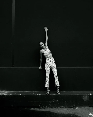 Monochrome shot of a solo adult female model in a zebra-print outfit leaning on a black wall with one arm raised above her head.