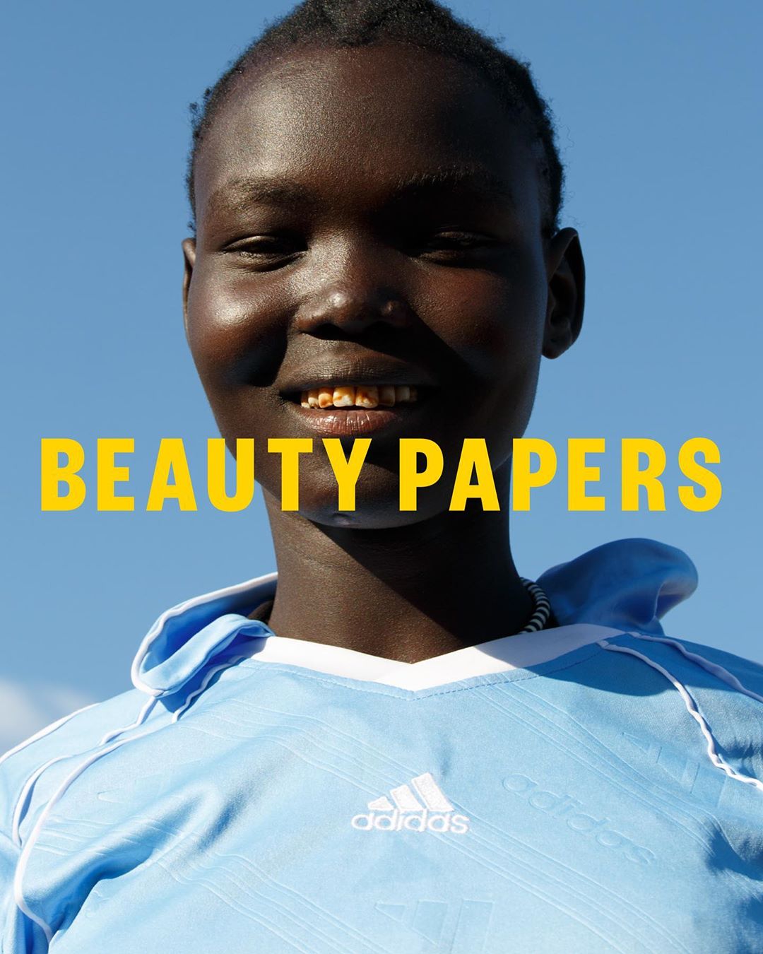 Portrait of a smiling, dark-skinned adult female with gold teeth wearing a light blue Adidas athletic jersey against a clear blue sky.