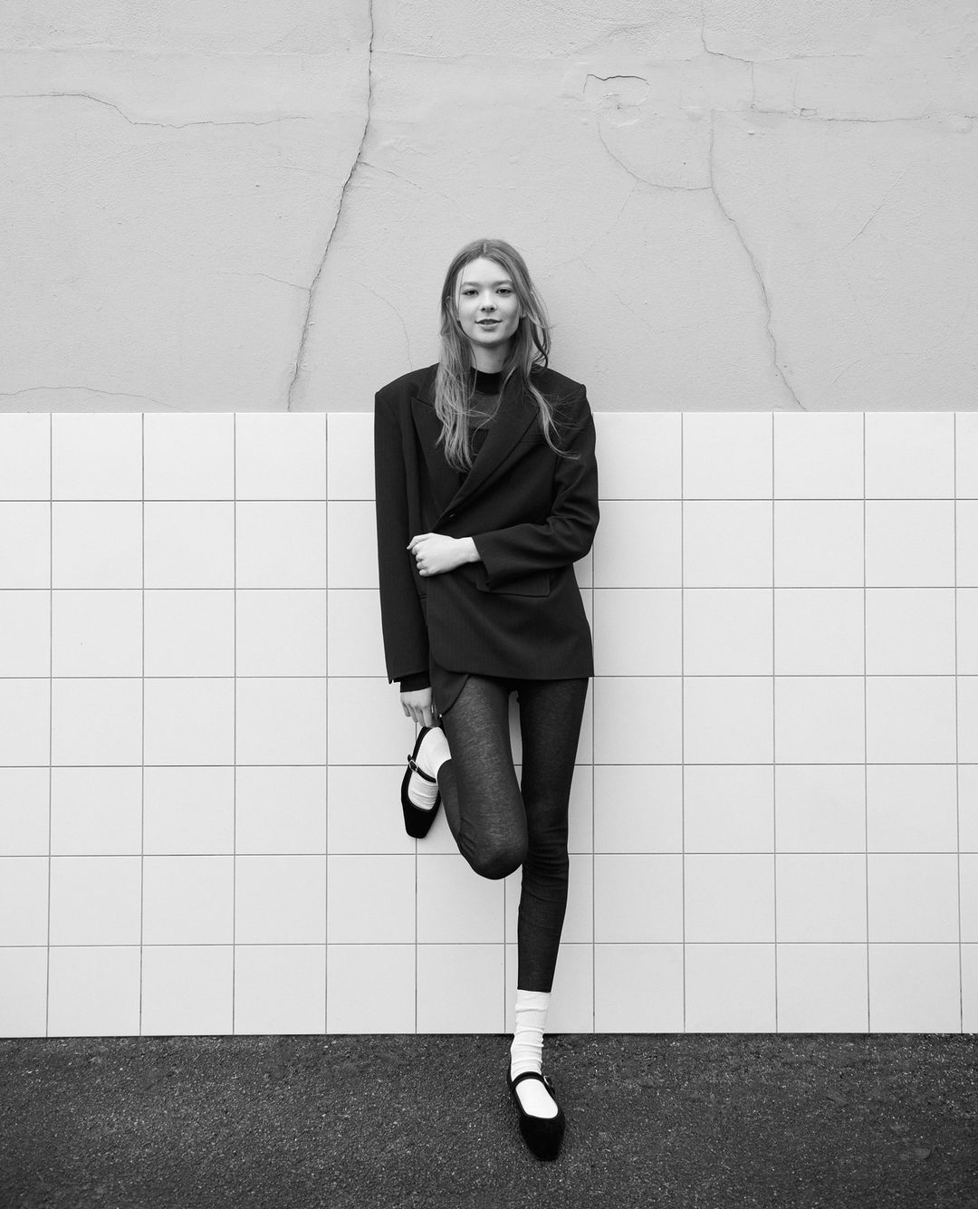 Black and white portrait of a young woman in an oversized blazer leaning against a tiled wall. Fashion-forward, stylish, and chic.