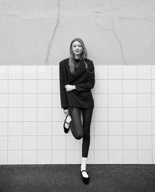 Black and white portrait of a young woman in an oversized blazer leaning against a tiled wall. Fashion-forward, stylish, and chic.