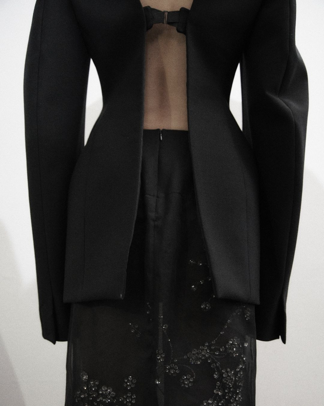 Formal black jacket and skirt ensemble features mesh panel and beaded design details, set against a bright studio backdrop.