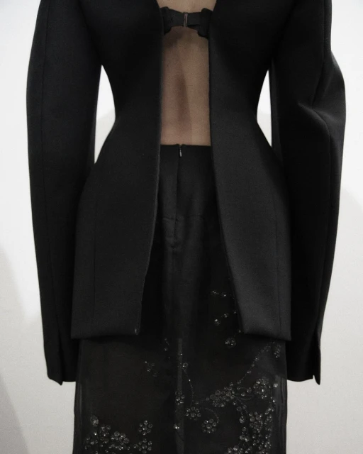 Formal black jacket and skirt ensemble features mesh panel and beaded design details, set against a bright studio backdrop.