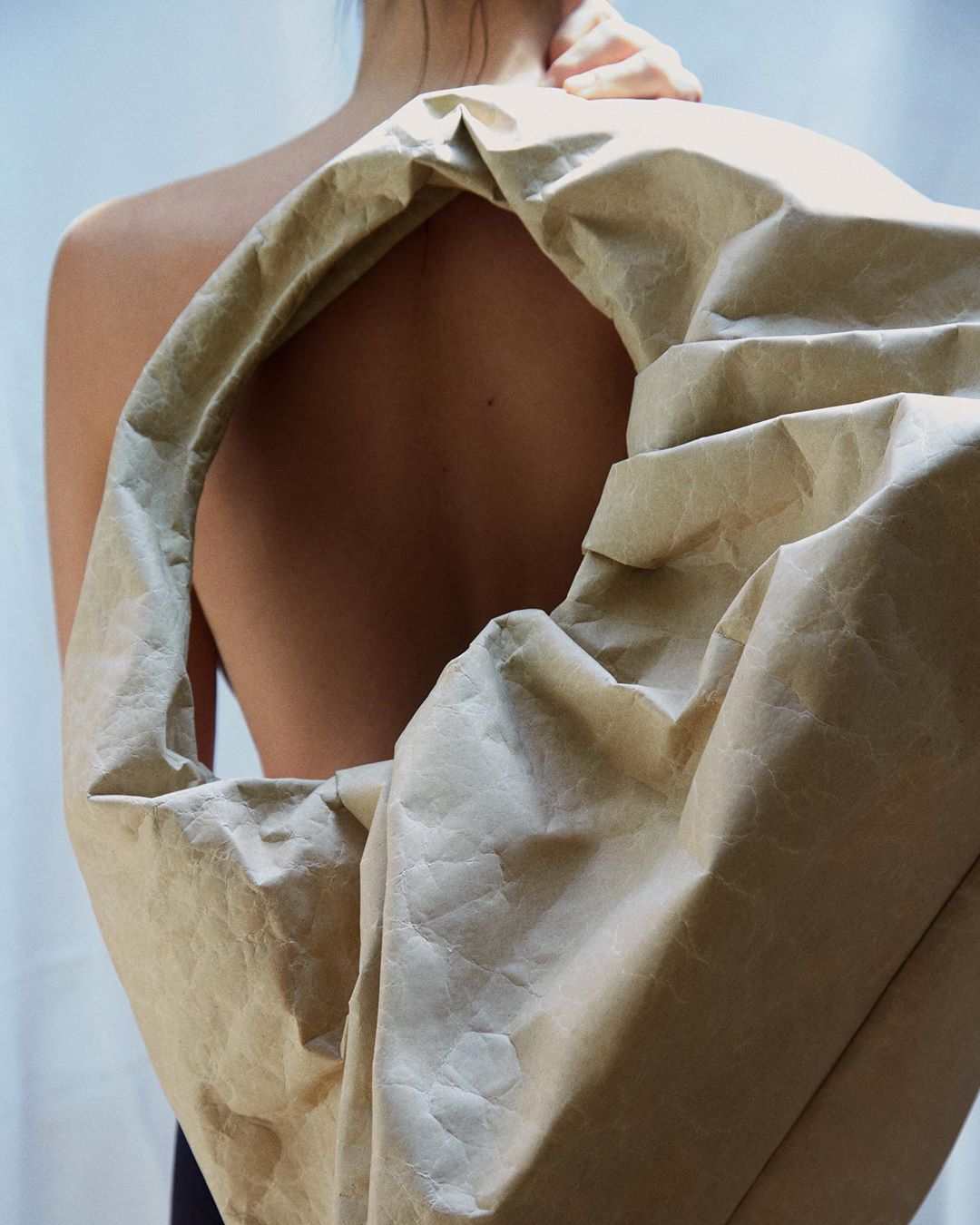 Back view of a partially visible woman holding a large beige paper bag over her shoulder against a solid light-grey background.