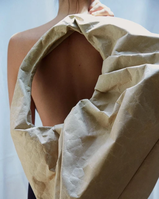 Back view of a partially visible woman holding a large beige paper bag over her shoulder against a solid light-grey background.