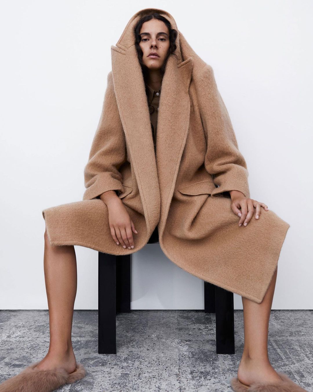 A young adult woman with dark curly hair sits on a black stool wearing a tan hooded wool coat and furry tan slippers in a photo studio.