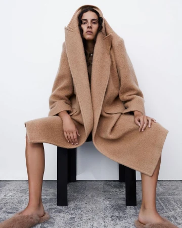 A young adult woman with dark curly hair sits on a black stool wearing a tan hooded wool coat and furry tan slippers in a photo studio.