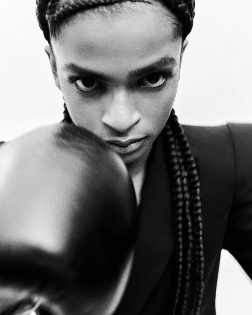 Monochrome portrait of a black woman with braided hair wearing a dark blazer and boxing glove staring directly at the viewer.