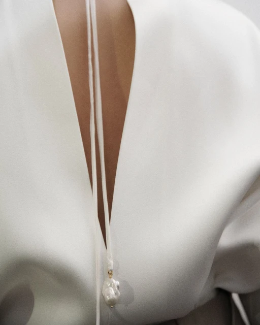 Close-up of a woman's back with white top and pearl pendant. The pendant hangs from thin white cords against the white top material.