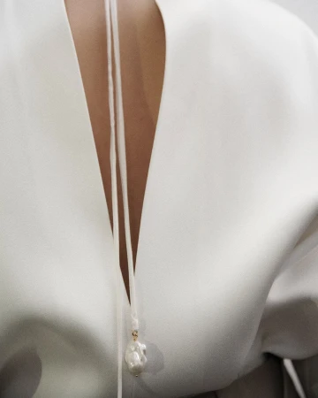 Close-up of a woman's back with white top and pearl pendant. The pendant hangs from thin white cords against the white top material.