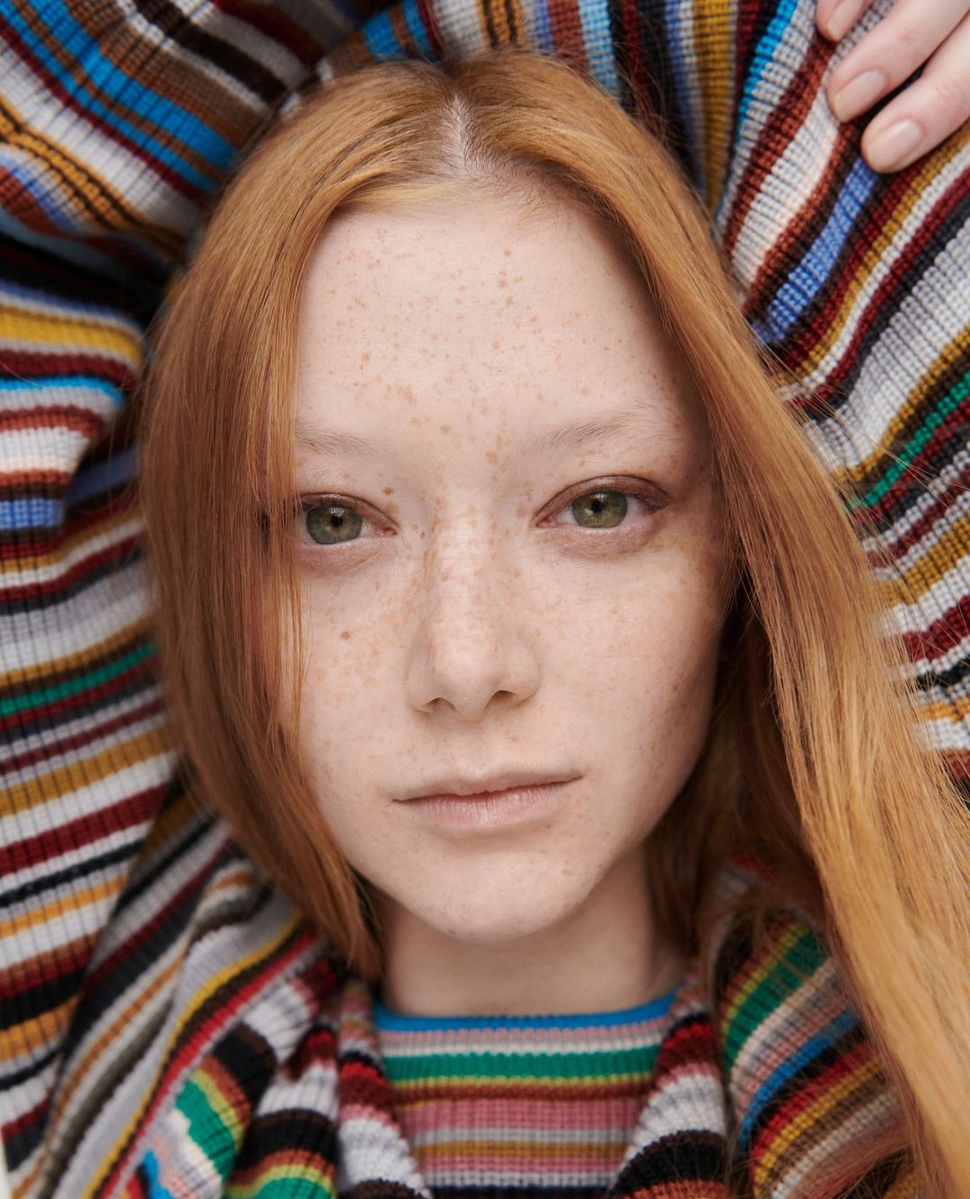 Close-up portrait of a young woman with red hair and freckles, wearing a striped top and matching jacket with vibrant multicolored stripes.