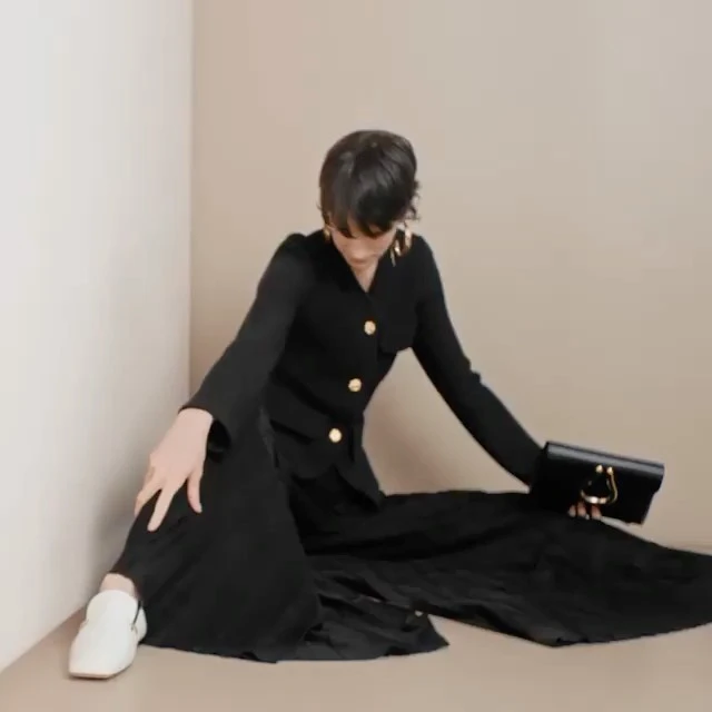 A woman in a black jacket, gold button details, wide-leg pants, and white shoes sits in a beige-toned corner, holding a black clutch.