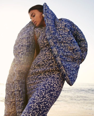 Model in blue and white patterned dress wearing a matching puffer coat against an outdoor sandy beach and ocean background.