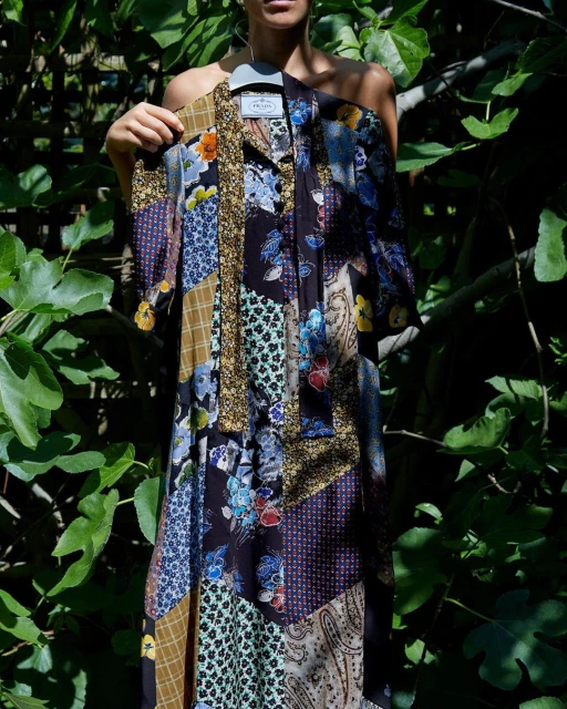 Woman holding a Prada patchwork print dress on a hanger in a lush green outdoor setting with dappled sunlight and visible foliage.