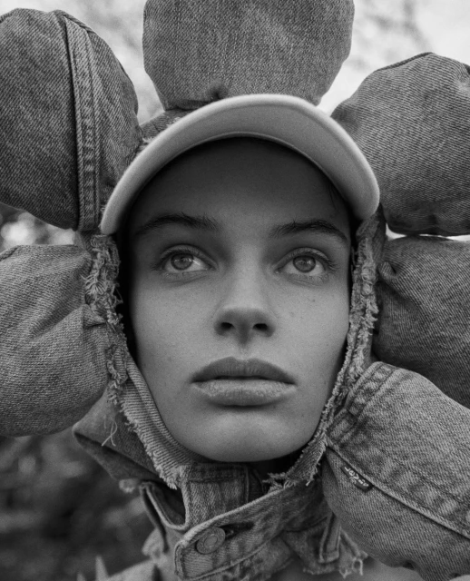 Black and white portrait of a woman wearing a denim hat with flower-like denim embellishments, photographed in a natural setting.