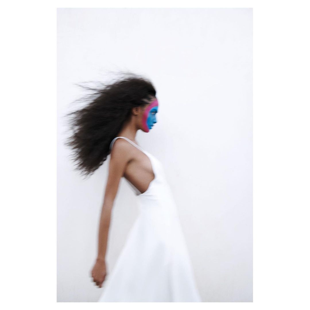 Woman in white dress with bold blue and pink face paint poses against a white background, with motion blur on her hair.