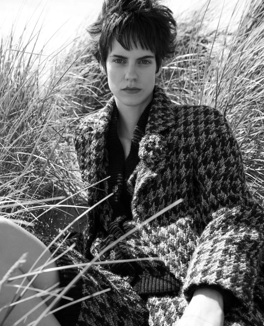 Black and white portrait of a woman with short dark hair wearing a patterned coat and dark top, sitting in tall grass outdoors.
