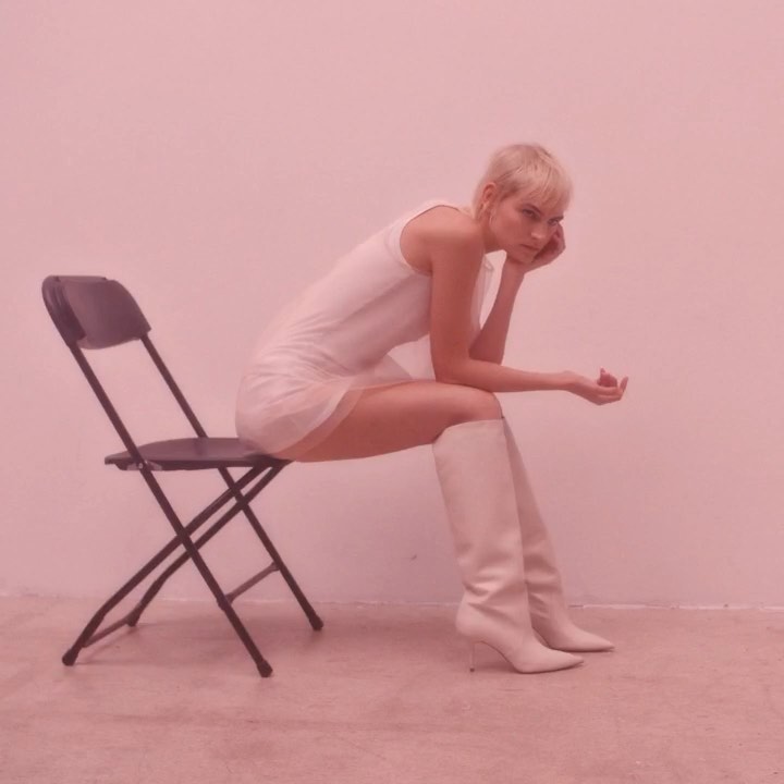 Woman with short blonde hair, wearing a white dress and boots, sitting on a black folding chair against a pink wall in a photo studio.